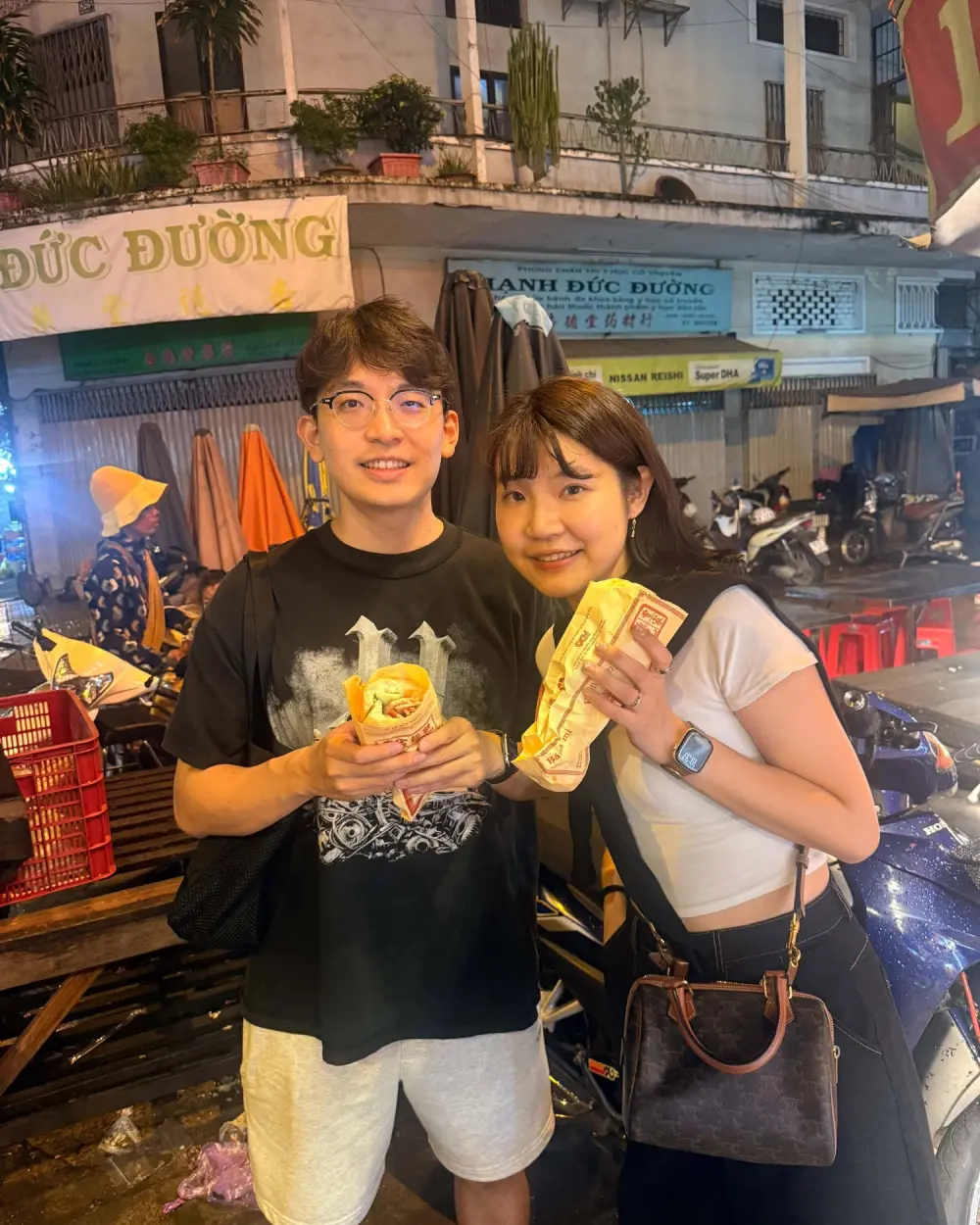 Two travelers holding fresh bánh mì on a night street food tour